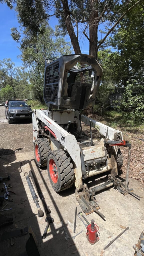 A white skid steer loader with its cab raised is parked outdoors on a concrete surface, surrounded by tools and parts, with trees and a car in the background.
