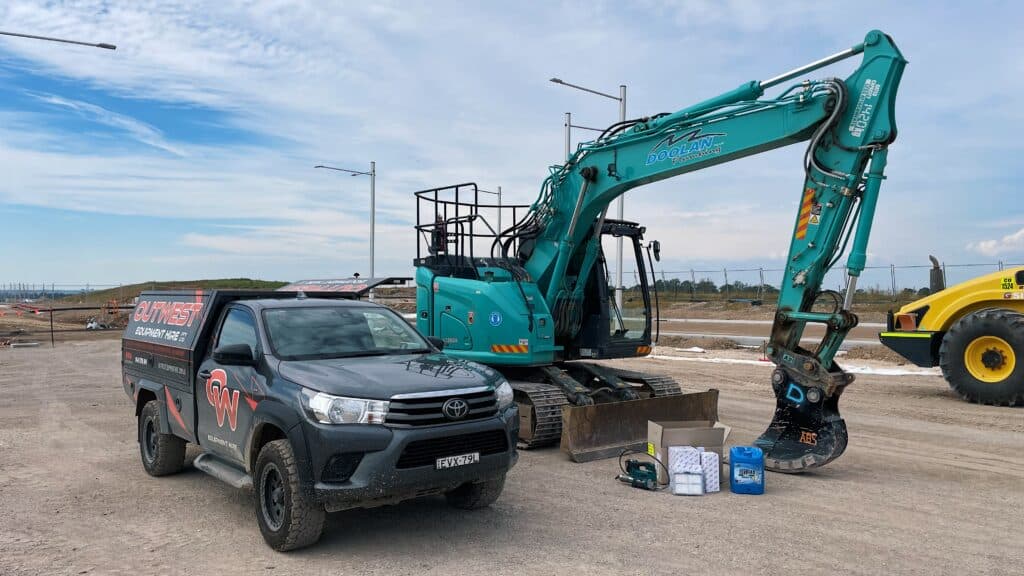 A teal excavator parked at a construction site beside a black utility lorry with company branding. Maintenance supplies are arranged on the ground in front.