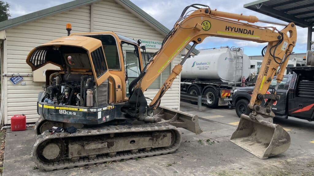 A yellow Hyundai excavator with its engine compartment open is parked on a concrete surface near a building and fuel lorry.