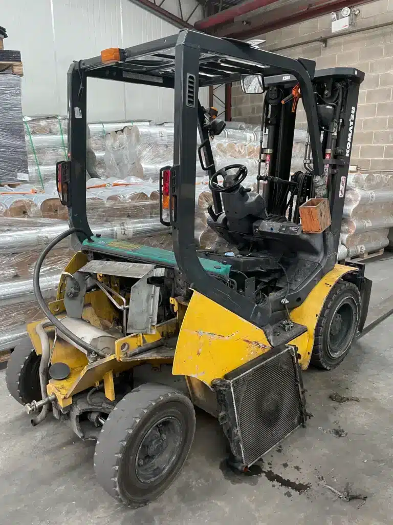 A yellow forklift with visible damage and missing rear panel is parked indoors near stacks of wrapped materials.