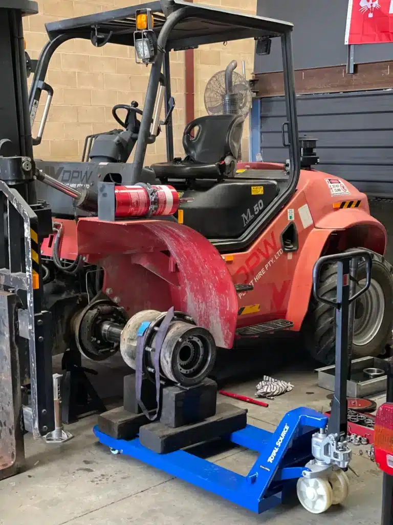 A red forklift is undergoing maintenance in a workshop, with a large mechanical component removed and placed on a blue trolley in front of it. Tools are scattered on the floor nearby.