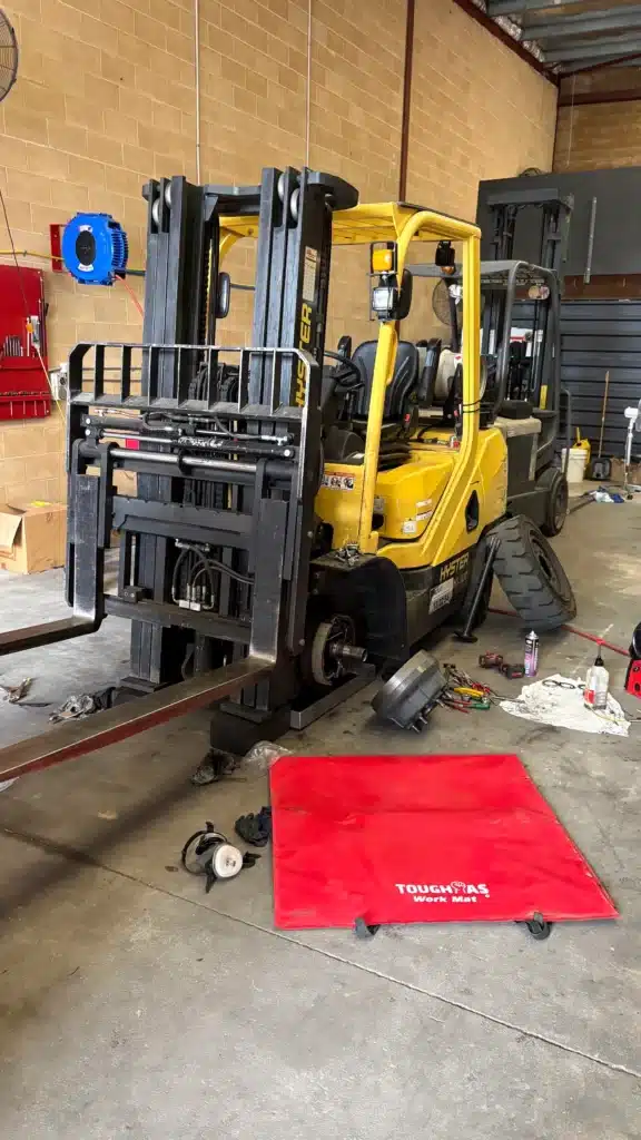 A yellow forklift is undergoing maintenance in a workshop; tools, parts, and a red Toughbuilt mat are scattered on the floor beside it.