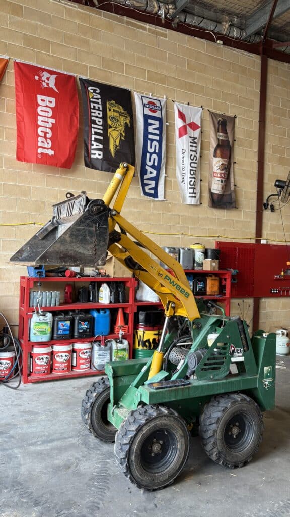 A small green and yellow skid steer loader with a raised bucket is parked indoors in a workshop with tools, supplies, and machinery banners on the wall behind it.