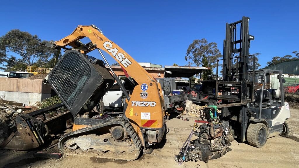 A CASE TR270 compact track loader is lifting a forklift, which is holding an engine at a construction yard under a clear blue sky.