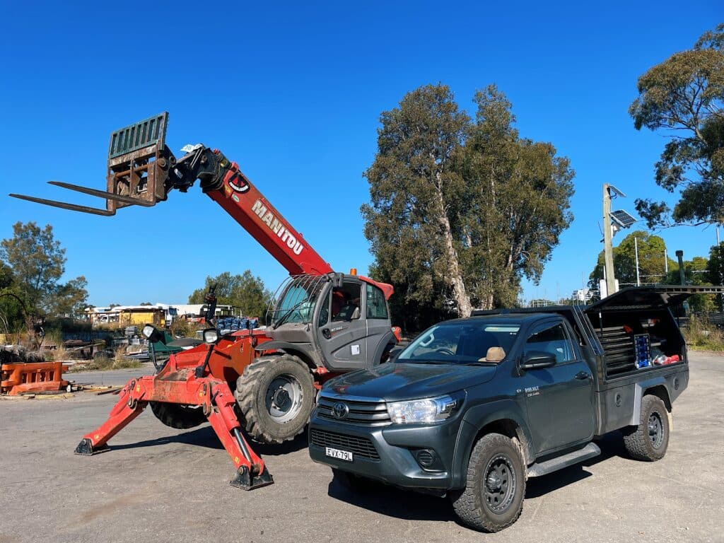 A red Manitou telehandler with raised forks is parked next to a grey utility lorry in an outdoor industrial area under a clear blue sky.