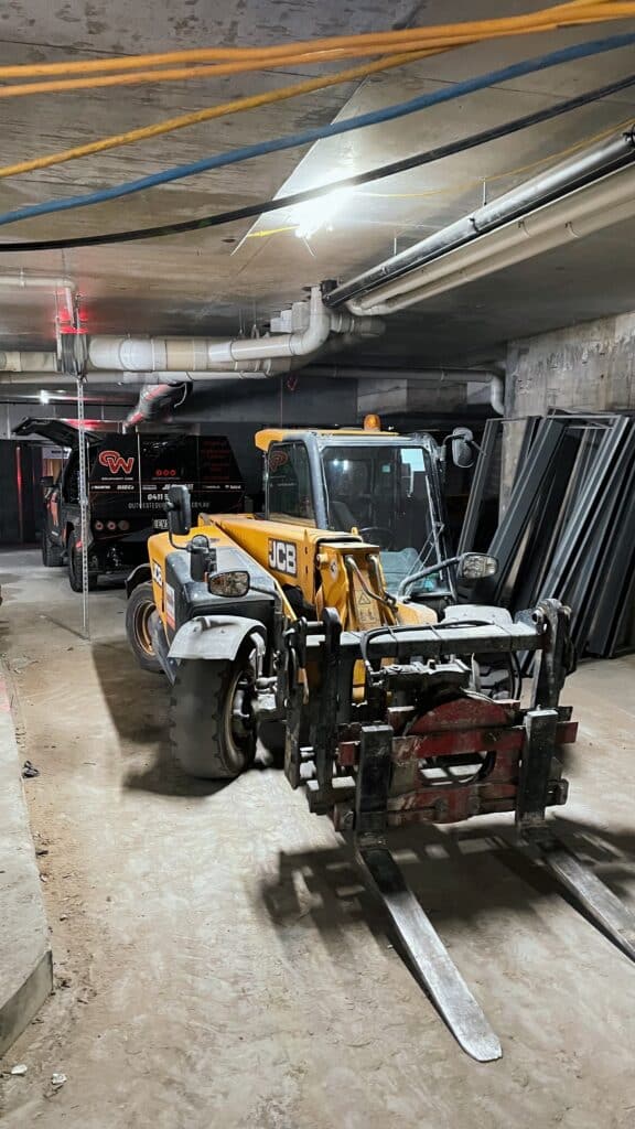 A yellow JCB telehandler forklift is parked indoors on a construction site with concrete walls and various building materials nearby.