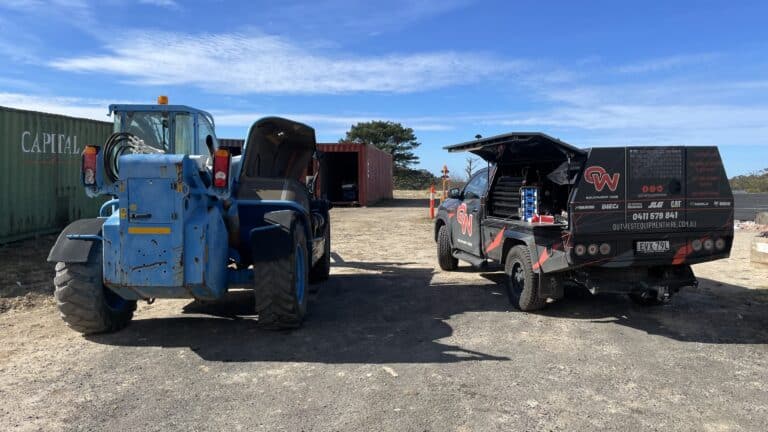 A blue tractor and a black utility vehicle with open bonnets are parked on a dirt lot, with shipping containers and a tree in the background under a blue sky.