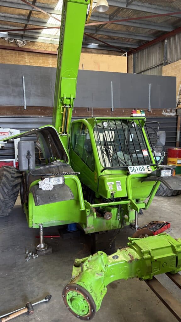 Bright green telehandler in a workshop with its front axle assembly removed, resting on stands. Tools and machinery are visible in the background.