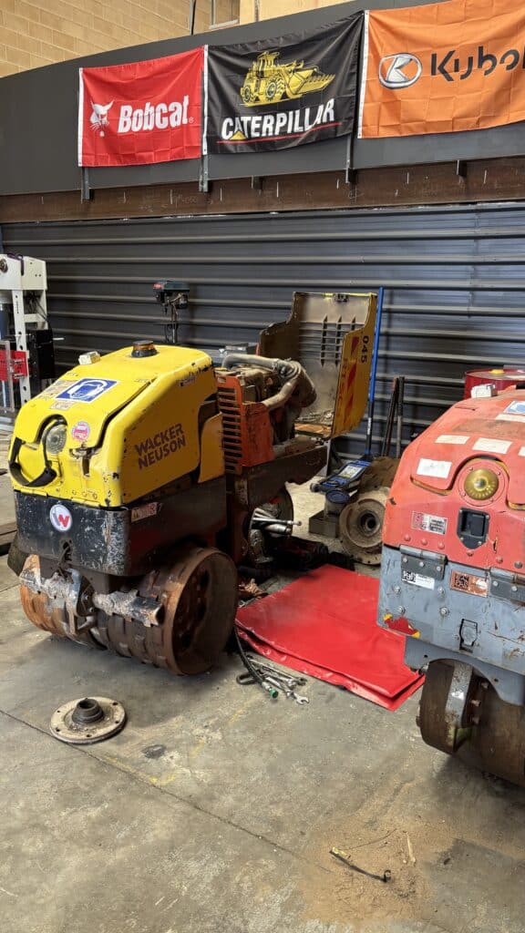 A partially dismantled Wacker Neuson compactor sits on a workshop floor next to another machine, with tools and parts scattered nearby.