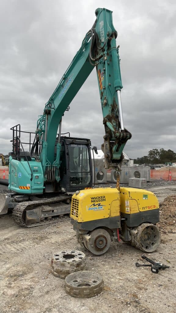 A large teal excavator moves or lifts a small yellow construction roller at a construction site with loose soil and scattered equipment.