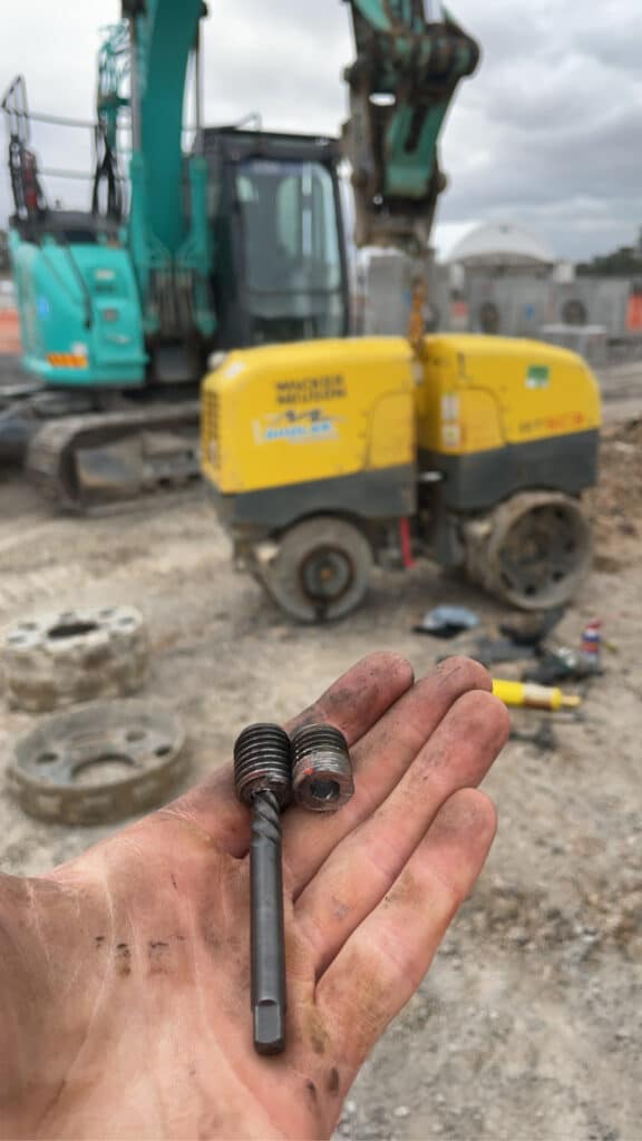 A hand holds two dirty, damaged bolts on a building site, with a digger and other equipment visible in the background.