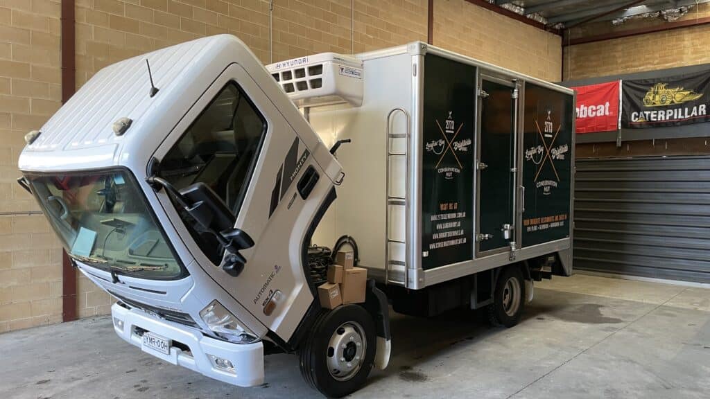A refrigerated lorry with its cab tilted forward is parked inside an industrial garage.